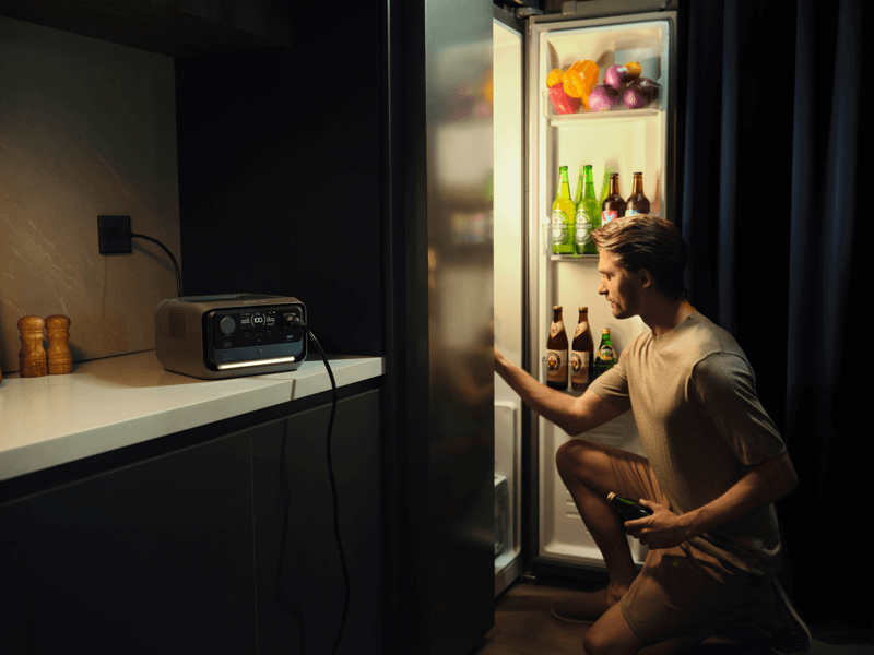 A person kneels by an open fridge filled with bottles and fresh produce, holding a beverage. An EcoFlow RIVER 3 Max power station sits on the nearby countertop, quietly humming with its X-Boost technology in the dimly lit room.