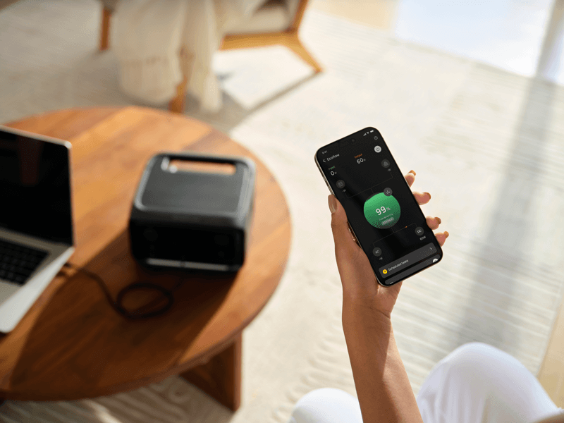 In a bright, modern living room, someone holds a smartphone displaying a 99% charge on an EcoFlow portable power station. The device sits on a wooden table next to a laptop, efficiently charging with its advanced X-Boost technology.