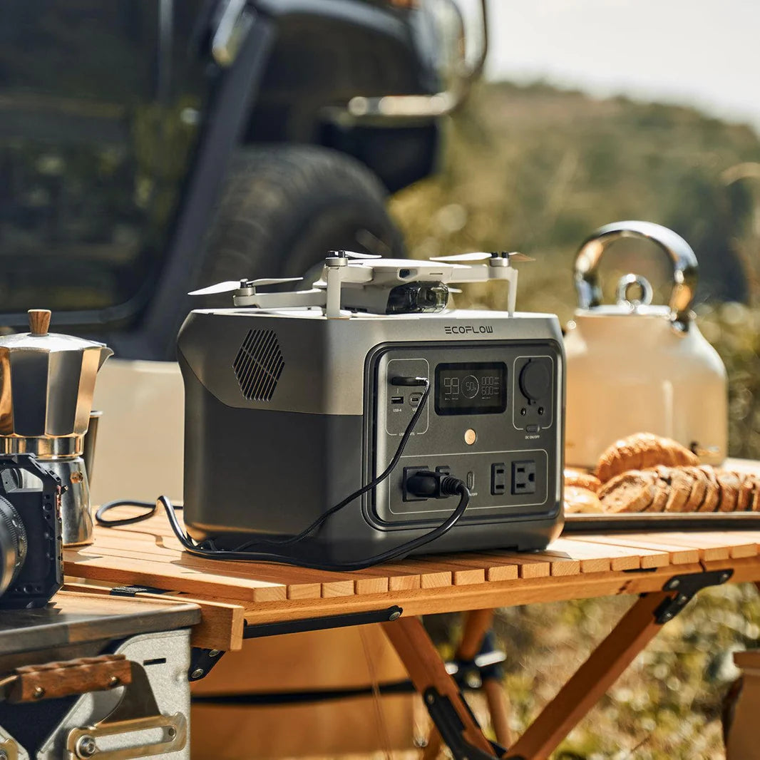 An EcoFlow portable power station sits on a wooden camping table outdoors, next to a coffee maker, bread, and kettle, with a vehicle and grassy landscape in the background—but the product description gives no details.
