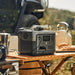 An EcoFlow portable power station sits on a wooden camping table outdoors, next to a coffee maker, bread, and kettle, with a vehicle and grassy landscape in the background—but the product description gives no details.