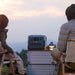 Two people sit outdoors on camping chairs facing an EcoFlow portable power station and lantern on a metal chest, with grass and a cloudy sky in the background.