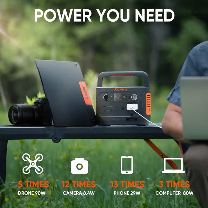 A Jackery portable power station sits on an outdoor table, charging a laptop with a camera nearby. Image text features its capabilities: charges a drone 5 times, camera 12 times, phone 13 times, and computer 3 times. A backdrop of blurred greenery completes the scene.