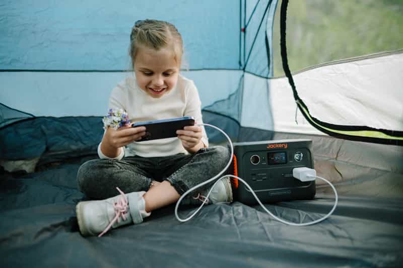 A child sits in a tent, holding a smartphone and smiling. Beside them is a Jackery portable power station with a device plugged in and charging. The tent's door is partially open, revealing greenery outside.