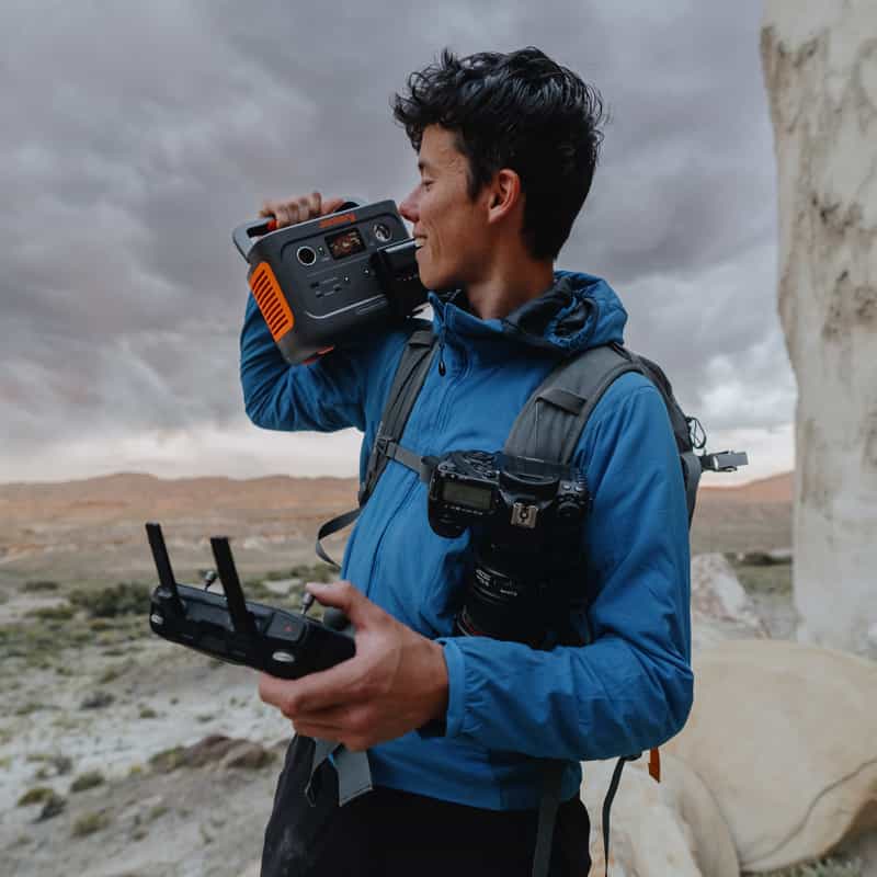 A person outdoors, wearing a blue jacket under a cloudy sky over rocky terrain, carries a Jackery portable power station on their shoulder and holds a drone controller. A camera is strapped to their chest.