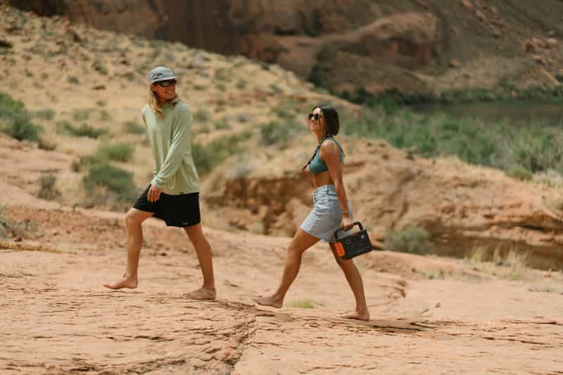 Two people walk barefoot on a rocky path in the desert. The person on the right carries a Jackery portable device. Both are casually dressed, surrounded by sparse vegetation with a body of water in the background.