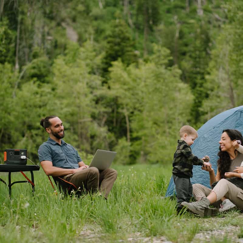 A man sits on a chair using a laptop beside a table with a Jackery device. Nearby, a woman and child laugh and play together on the grass in front of their tent, surrounded by lush green trees.