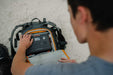 Outdoors, someone in a gray shirt is putting a Jackery portable power station into the open compartment of their backpack on a textured surface.