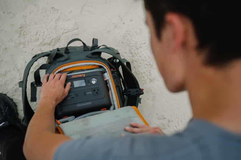 Outdoors, someone in a gray shirt is putting a Jackery portable power station into the open compartment of their backpack on a textured surface.