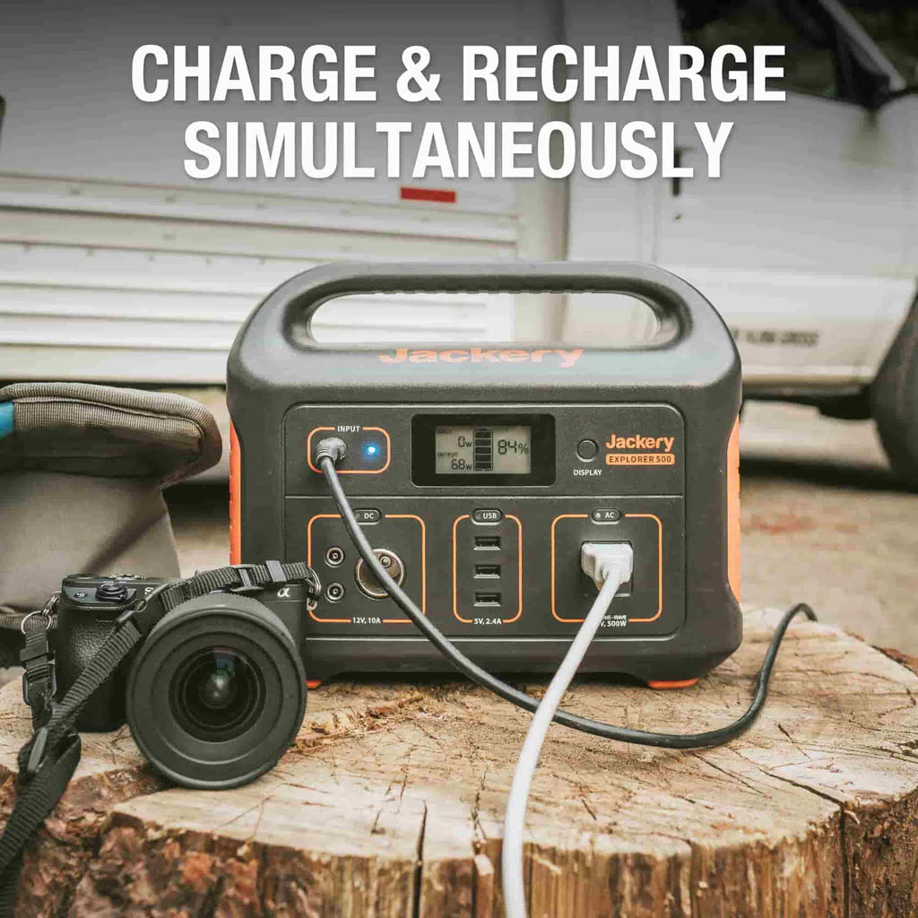 On a tree stump outdoors, a Jackery portable power station with high battery capacity is shown powering a camera and another device through its multiple charging ports. The text above states "Charge & Recharge Simultaneously," with a vehicle visible in the background.