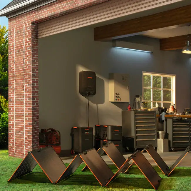 Solar panels are set up in front of an open garage equipped for a home backup system featuring Jackery's LiFePO4 battery. Inside, toolboxes, a workbench, and wall-mounted equipment are ready. The green grass and brick walls enhance the scene's eco-friendly vibe.