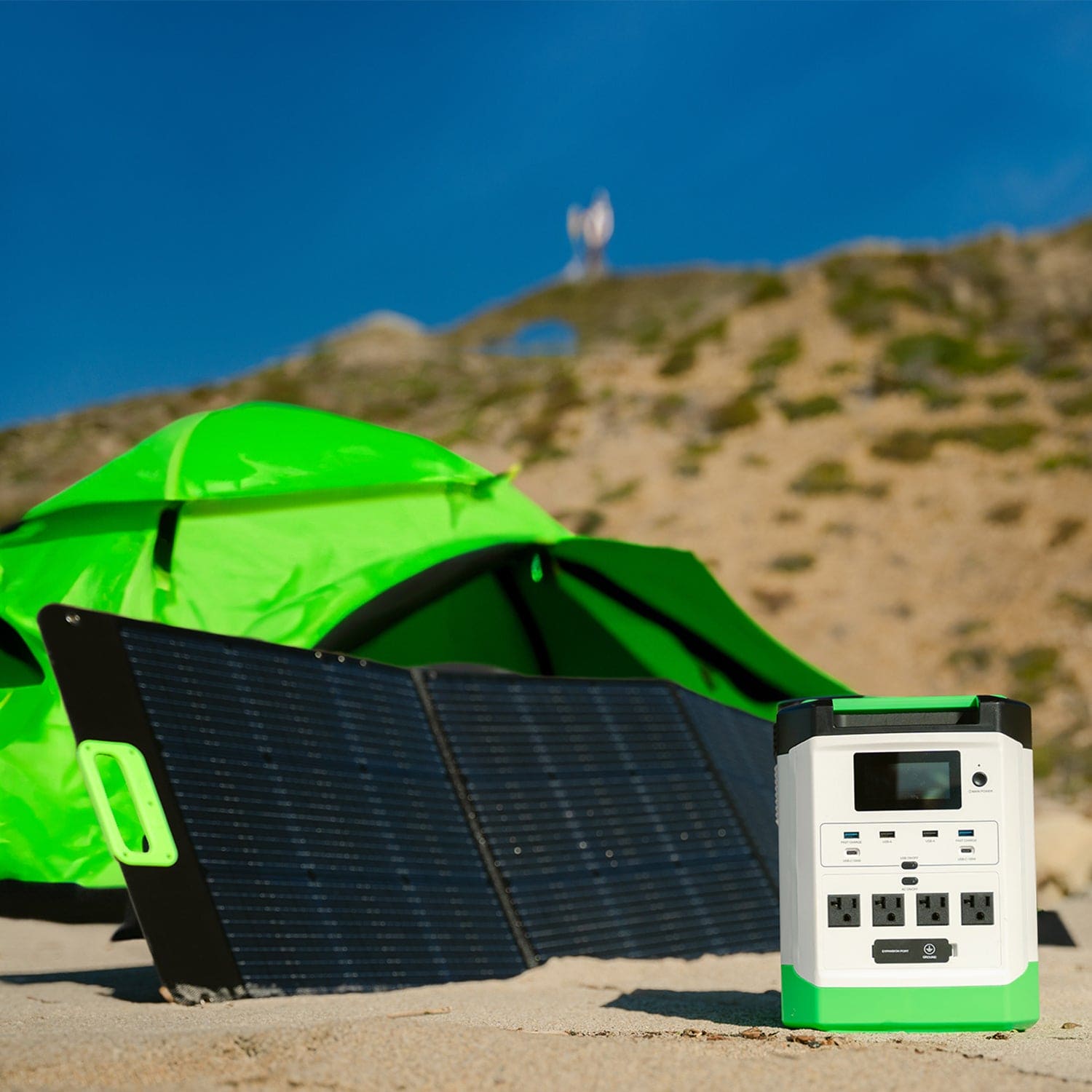 A Nature's Generator portable power station with a LiFePO4 battery and foldable solar panel sit on sand in front of a bright green tent, with grassy hills and a blurred person visible in the background.