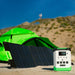 A Nature's Generator portable power station with a LiFePO4 battery and foldable solar panel sit on sand in front of a bright green tent, with grassy hills and a blurred person visible in the background.