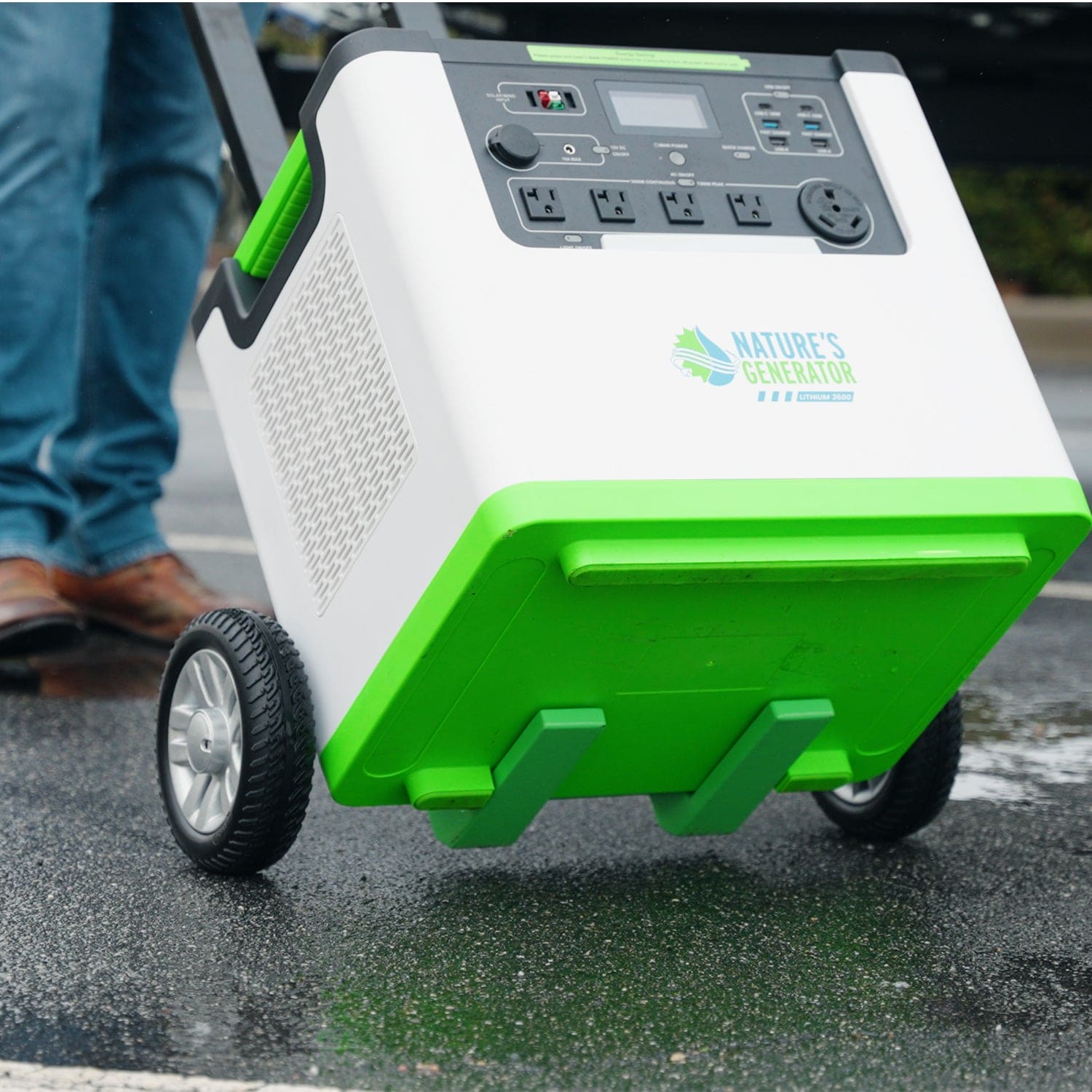 A Nature's Generator portable Lithium 3600 power station with wheels is being rolled on wet pavement. Its control panel and multiple outlets are visible, with a person's feet in jeans and shoes partially seen in the background.