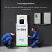 Two technicians in blue uniforms install a Nature's Generator expandable energy system. One crouches while the other adjusts the unit, with electrical panels on the wall and instructions posted above them.