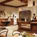 A barista pours a drink behind a pastry case in a cozy café with wood accents. The menu board and coffee machine, powered by Nature's Generator LiFePO4 battery, are visible as a man sits alone at a table using his phone.