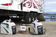 Two quiet portable generators, including a Pulsar inverter generator, and a power box are connected by cables beside a parked RV on sandy ground near water, with inflatable watercraft visible in the background.