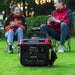 A red and black PowerSmart portable generator sits on the grass, supplying camping power, as two people in jackets relax with mugs on foldable chairs in front of lush greenery.