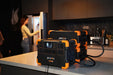 On a kitchen counter, several Pecron E2000LFP power stations are neatly stacked and interconnected with cables. In the background, a person wearing a yellow shirt and jeans is opening the refrigerator. Surrounded by dark cabinets and various items, these power stations feature an expandable capacity of up to 8064Wh and include a 2000W Pure Sine Wave Inverter.