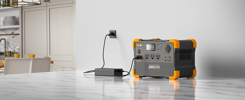 On a marble countertop, a Pecron portable power station with orange accents is showcased. It features fast charging technology and an E600LFP LiFePO4 Battery. A device is connected to it via a charging cable, and it sits near a white wall with a power outlet, while kitchen cabinets and dishware blur in the background.