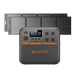 A Bluetti portable power station, branded with the Bluetti name, is positioned in front of four solar panels. The power station features multiple outlets and an LED display, and is mainly black with orange accents. The background is a plain white.