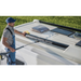 Wearing a cap and overalls, someone operates a Renogy power washer to clean the roof of a white RV equipped with solar panels and a skylight, set against a grassy backdrop.