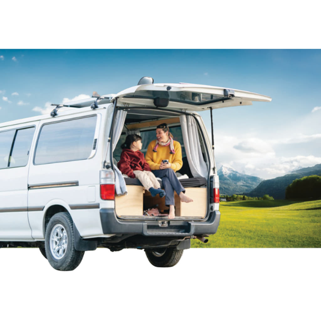 Two people relax in the back of a white Renogy van with its rear door open, parked on a grassy patch. They are holding beverages and seem to be enjoying a conversation amidst the scenic landscape featuring mountains and a blue sky.