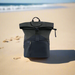 An EcoFlow RIVER 2 Travel Bag in black, featuring a roll-top design and zipper pockets, rests on sandy beach with the ocean and shoreline in the background.