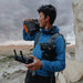 A person in a blue jacket holds a Jackery portable power station on their shoulder and a drone controller in the other hand, highlighting features like Smart App Control. With a camera strapped to their chest, they stand against a rugged landscape beneath a cloudy sky.