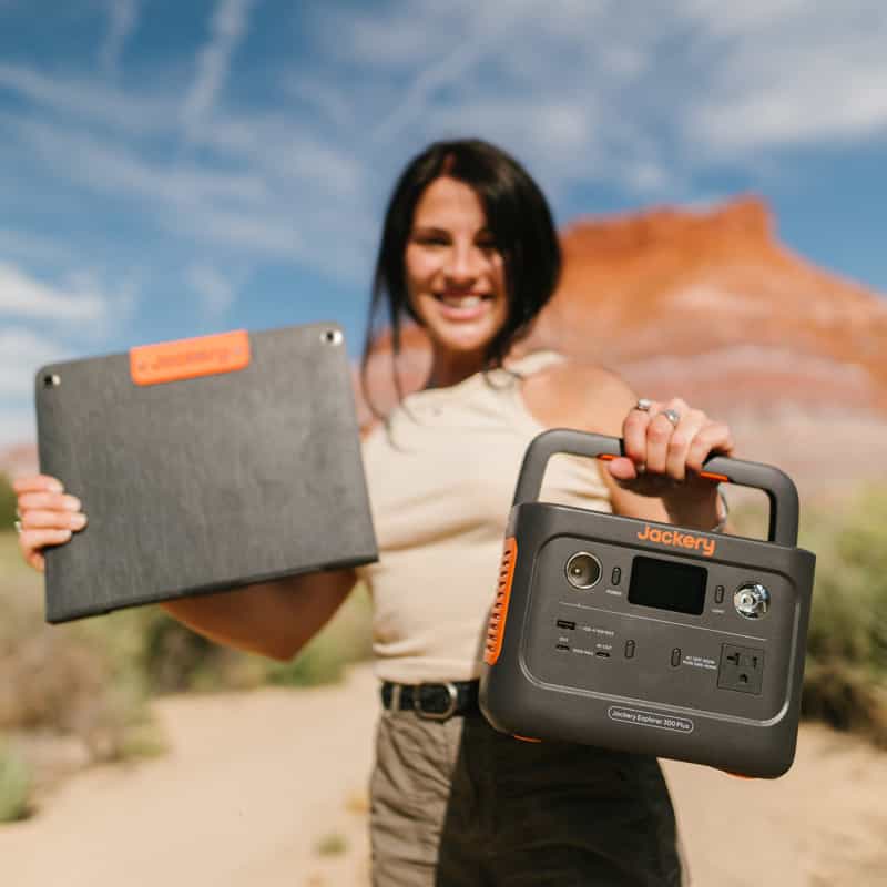 A woman proudly holds the Jackery Solar Generator and a solar panel amid red rock formations, enjoying a blue sky with scattered clouds and utilizing ultra-fast charging for her adventures.