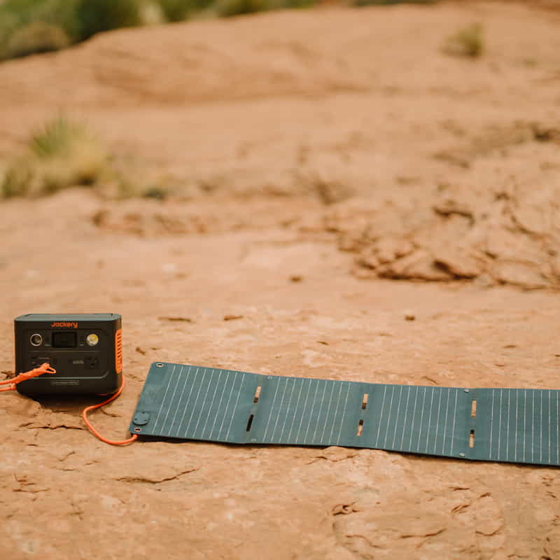 A Jackery Solar Generator sits on rocky terrain with an unfolded solar panel linked to a compact power station by an orange cable. It efficiently harnesses solar energy with ultra-fast charging against a backdrop of blurred nature.