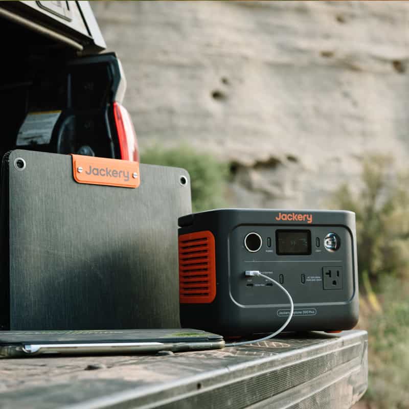 A Jackery portable power station, featuring an orange and gray design with visible USB ports and outlets for ultra-fast charging, sits on the back of a truck. Nearby, a Jackery Solar Generator panel is propped against a rocky landscape.