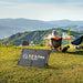 Two people sit in camping chairs at a table on grass with mountains in the background. A portable solar panel and generator offer convenient off-grid power in the foreground. A dog sits near the chairs, and various camping items are scattered across the table.