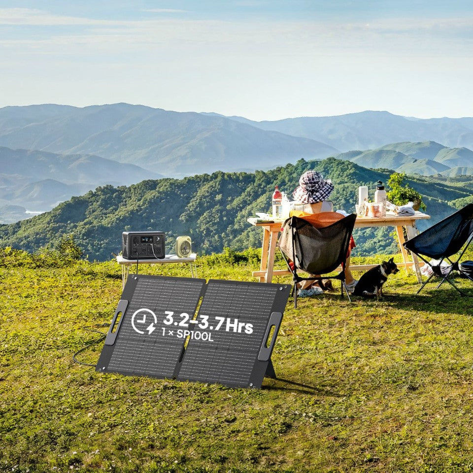 Two people sit in camping chairs at a table on grass with mountains in the background. A portable solar panel and generator offer convenient off-grid power in the foreground. A dog sits near the chairs, and various camping items are scattered across the table.