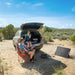 Two people relax at a campsite next to an SUV with its trunk open. One sits comfortably in a chair with a laptop, while the other enjoys a drink from the back of the vehicle. A portable monocrystalline solar panel and power station are set up on the ground nearby, providing reliable off-grid power for their outdoor adventure.