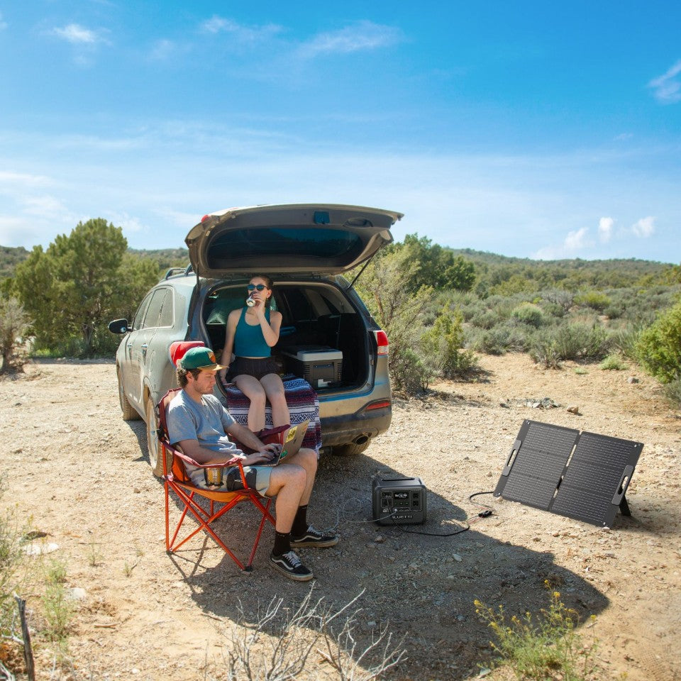 Two people relax at a campsite next to an SUV with its trunk open. One sits comfortably in a chair with a laptop, while the other enjoys a drink from the back of the vehicle. A portable monocrystalline solar panel and power station are set up on the ground nearby, providing reliable off-grid power for their outdoor adventure.