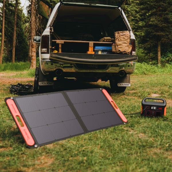 A Jackery SolarSaga 100W Solar Panel is set up on the grass near a parked truck’s open tailgate, alongside camping gear and a power station, showcasing a portable energy solution with trees in the background.