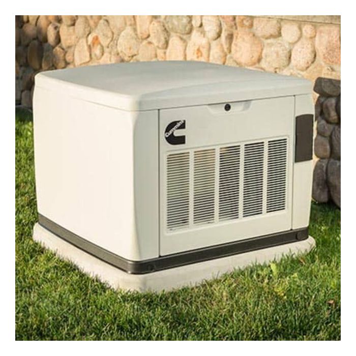 A Cummins white outdoor generator with a ventilation grill rests on a sturdy Genpad 3 Inch Concrete Composite Mounting Pad, nestled on grassy ground beside a stone wall.