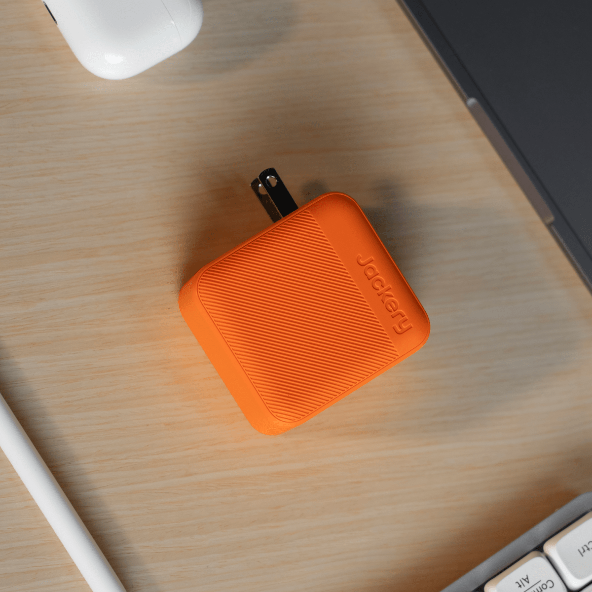 A compact orange Jackery USB-C charger with foldable prongs sits on a wooden desk beside a laptop, white stylus, keyboard, and part of a white device.