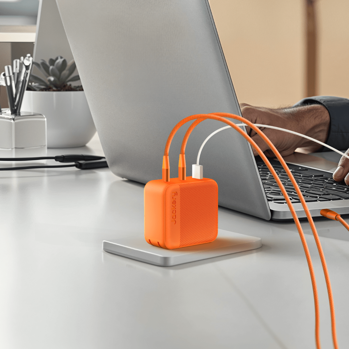 A Jackery orange 100W fast charging hub with multiple cords connects to a laptop on a white desk, as a person’s hand rests on the keyboard. A potted plant and notebook sit in the background.