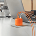 A Jackery orange 100W fast charging hub with multiple cords connects to a laptop on a white desk, as a person’s hand rests on the keyboard. A potted plant and notebook sit in the background.