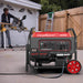 A PowerSmart red and black dual fuel generator with control panel and wheels is in the foreground, while a person uses a power saw near a white garage in the background. A wooden fence and ladder are also visible.
