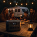 An EcoFlow portable power station sits on a wooden picnic table next to a glowing lantern as a family of four enjoys dinner outdoors near their camper, with string lights above adding to the cozy evening atmosphere.