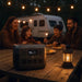 An EcoFlow portable power station sits on a wooden picnic table next to a glowing lantern as a family of four enjoys dinner outdoors near their camper, with string lights above adding to the cozy evening atmosphere.