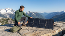 A hiker kneeling on a granite ledge high on a mountain ridge beside a Jackery 100Wair foldable solar panel, its four dark monocrystalline segments angled to the morning sun and a cable running to a backpack, snow-capped peaks behind.