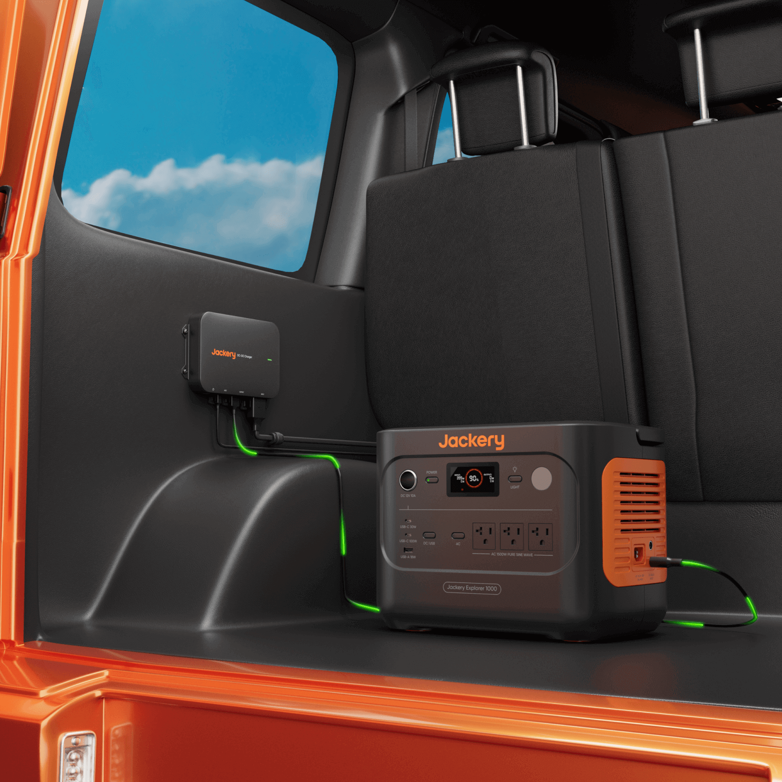 A Jackery portable power station and charging accessory, including a 600W vehicle charger, are connected inside the rear of an orange vehicle with cables plugged in. The sky is visible through the window. Brand: Jackery.