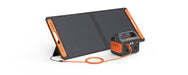 A Jackery foldable solar panel connects to a portable power station with an orange DC extension cable. Both black devices feature orange accents and display the "Jackery" brand name.