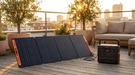 A Jackery E1500Ultra portable power station on a city rooftop terrace at golden hour, connected by cable to a Jackery SolarSaga 200W foldable solar panel standing open on its kickstand, with olive trees and the skyline behind.