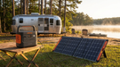 A Jackery Explorer 2000 v2 portable power station on a folding table at a lakeside campsite, cabled to a large Jackery foldable solar panel on the grass, with a silver travel trailer, camp chairs and mist on the water at golden hour.