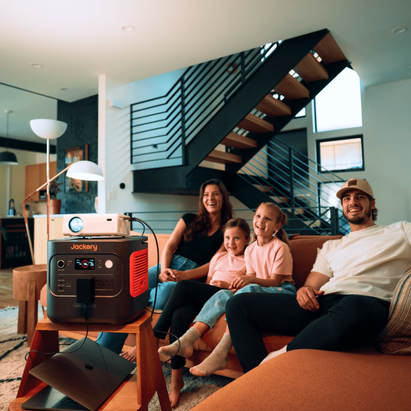 A family of four sits on a living room couch, smiling as they watch a projector movie powered by a Jackery portable power station placed on a small table in front of them. A staircase appears in the background.