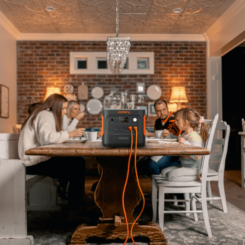 A family of five sits around a wooden dining table in a cozy room with brick walls, while a Jackery LiFePO₄ generator with bright orange cables rests on the table in the foreground.